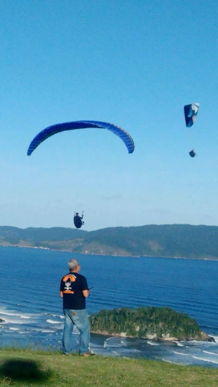 Na beira da rampa do Morro do Itararé, um instrutor acompanha parapentes voando sobre o mar e a ilha