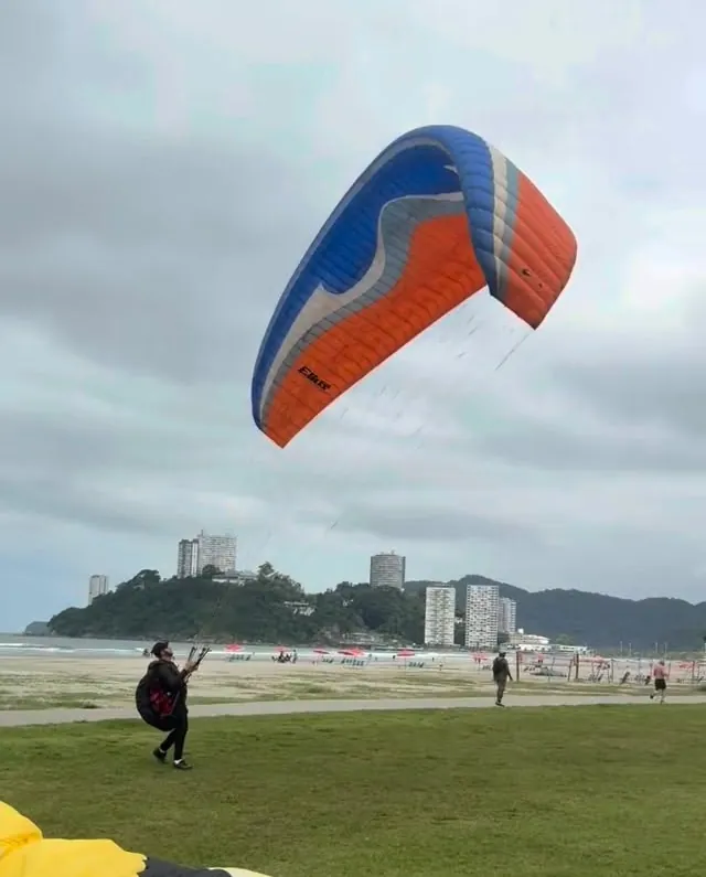 Aluna controlando uma vela laranja e azul na praia, com o morro ao fundo